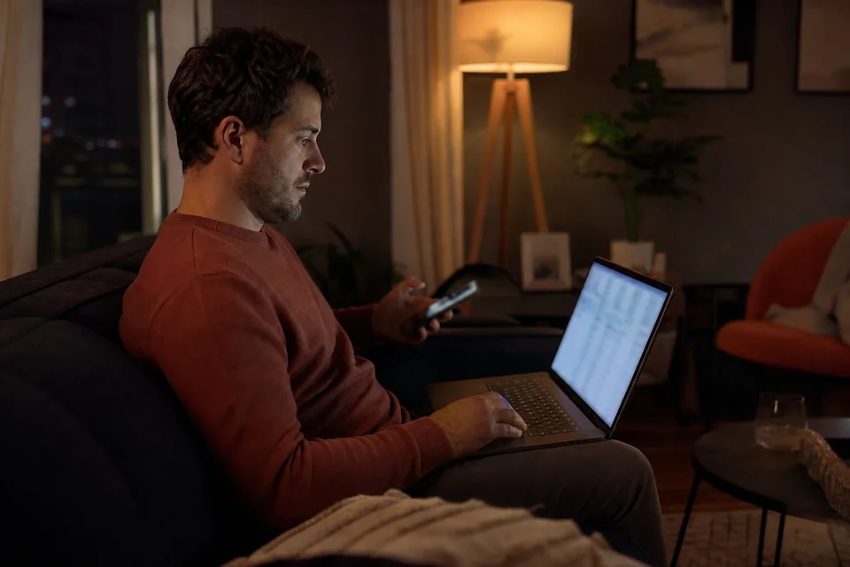 Man on a sofa at night going through a long list of video purchase receipts on his laptop