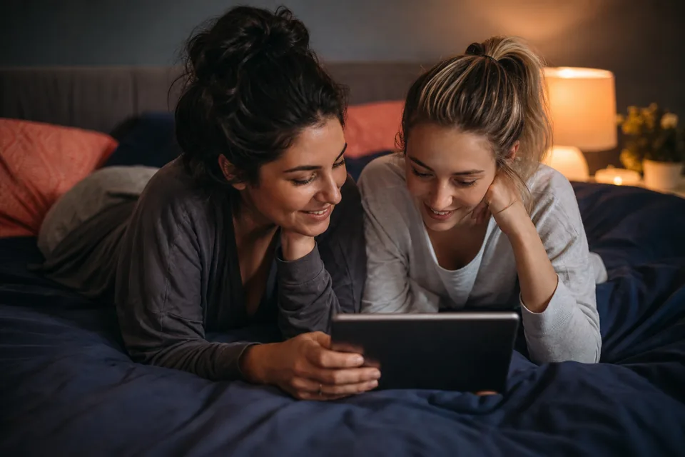Two women on a bed in the evening choosing what to watch together on a tablet